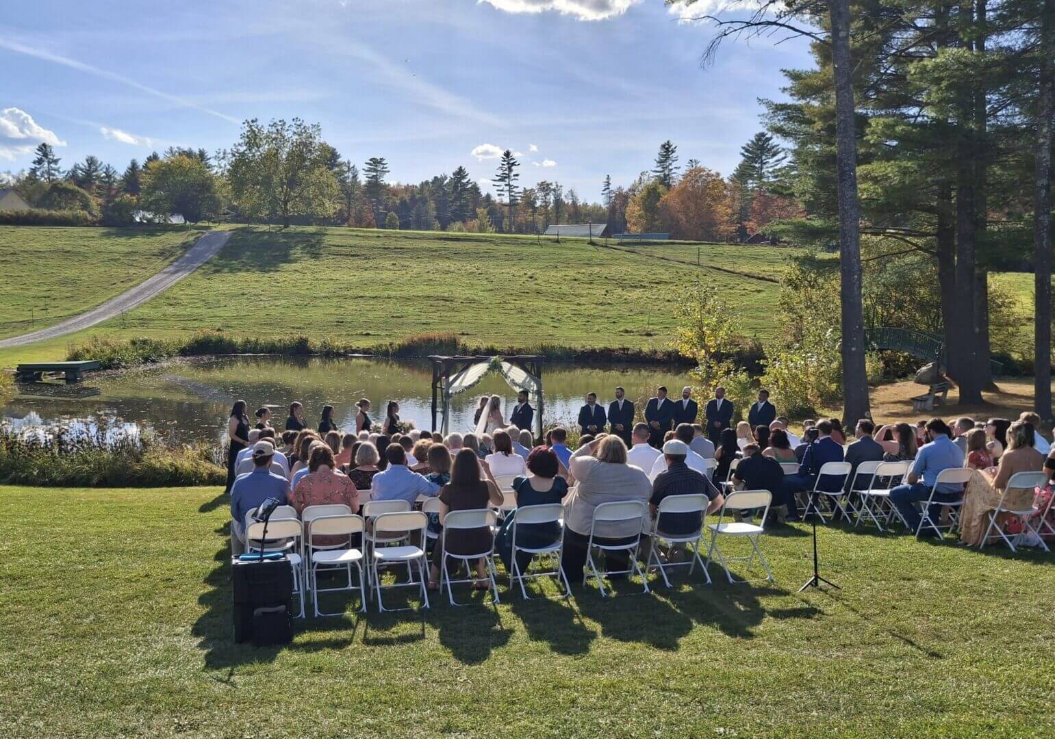 Outdoor wedding ceremony with guests seated on chairs under a partly cloudy sky.